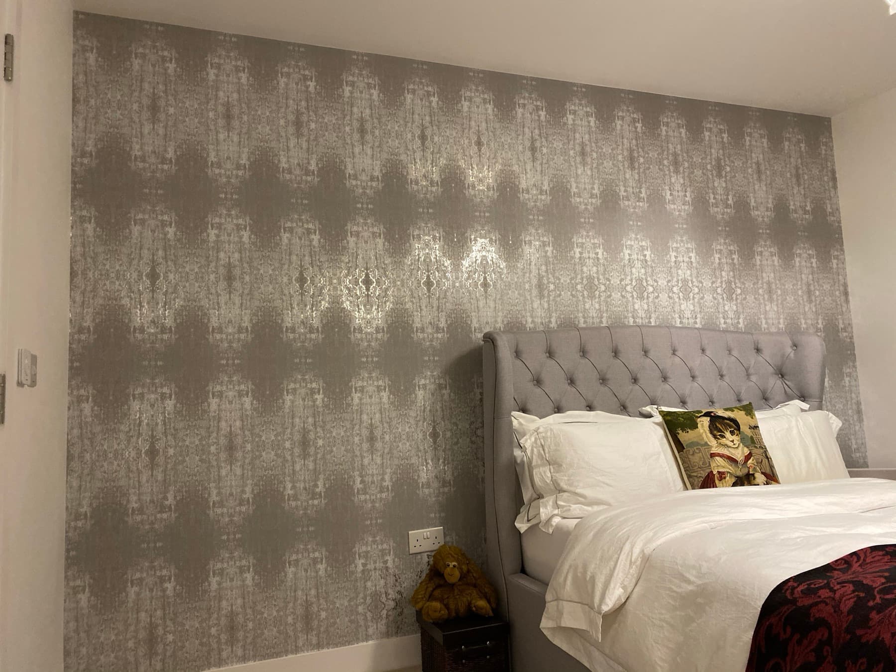 Bedroom with a silver damask wallpaper behind a buttoned headboard, pattern matched across the full wall