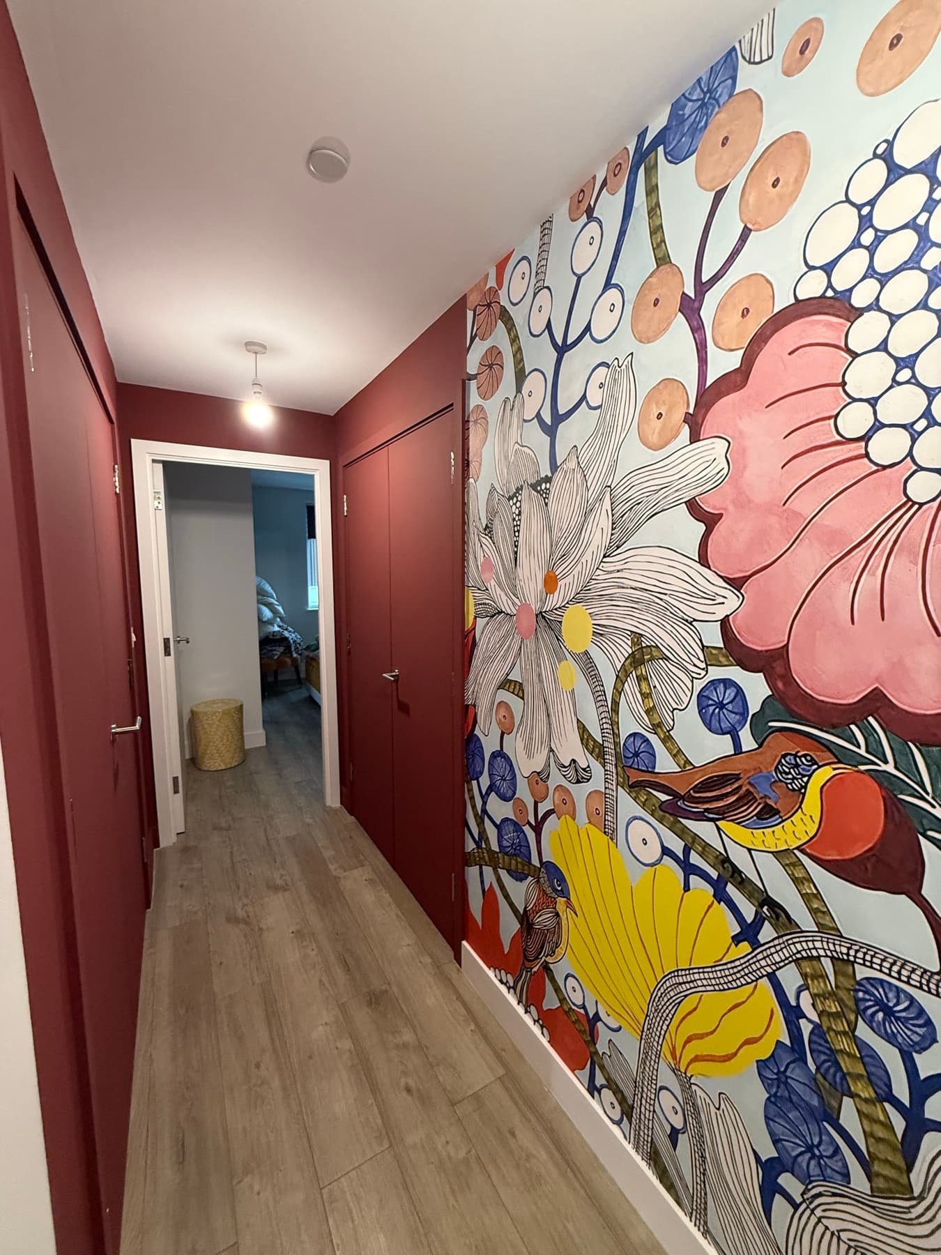 Narrow entrance hall with a large-scale floral and bird mural wallpaper down one wall, oxblood painted doors opposite and an oak-effect floor